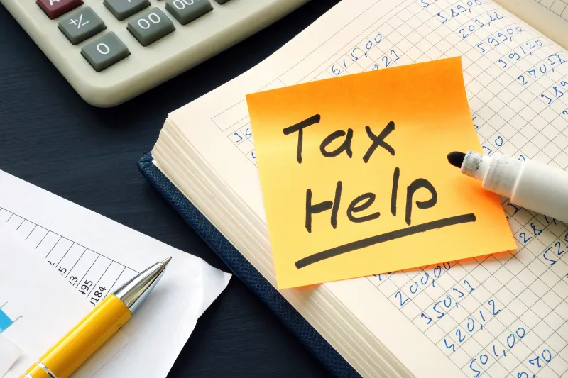 Yellow post-it note with the worth Tax Help written in black, resting on a grid notebook with a calculator also visible.