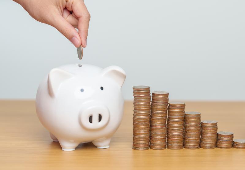Someone putting a coin into a piggy bank on a table beside piles of more coins